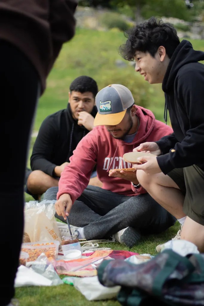 Male students sit on a picnic blanket and help themselves to sandwich ingredients.