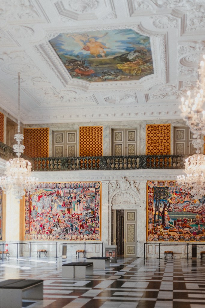 The Great Hall inside Christiansborg Palace, featuring an ornate white ceiling and two grand chandeliers.